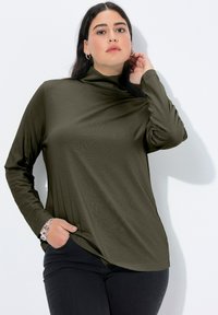 Tmavě zelený top s dlouhým rukávem a vysokým límcem, měkká textura a volný střih, kombinovaný s černými džínami; modelka má na sobě stříbrný náramek.