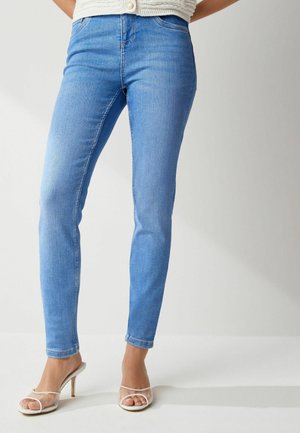 Femme portant un jean skinny bleu clair et des sandales blanches à talons hauts, debout sur un fond uni.
