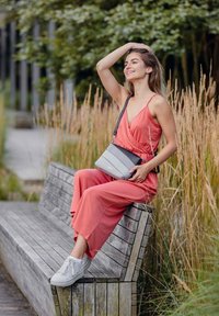 Šedá crossbody taška s texturovanými pruhy, nošená s korálovým overalem, v kombinaci s bílými teniskami, sedící na dřevěné lavici.