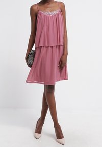 Rosa, schichtendes Kleid mit einem runden Ausschnitt, verziert mit silbernen Verzierungen. Glattes Material und eine fließende Silhouette. Einfache High-Heels.