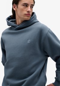 Felpa con cappuccio in un morbido tessuto grigio-blu. Presenta una vestibilità rilassata, tasca a marsupio e un logo ricamato discreto sul petto.