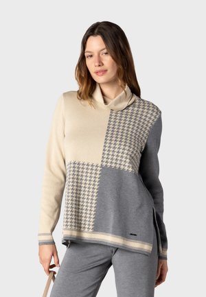 Kvinde iført en beige og grå hundetandmønstret rullekravesweater og grå bukser, der holder en beige strop med sin venstre hånd.