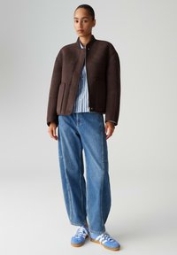 Giacca bomber marrone con texture liscia e collo a coste, abbinata a una camicia a righe azzurra chiara e jeans blu larghi. Completa il look un paio di sneakers blu.