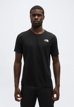 Nuori mies seisoo kuvattuna eteenpäin päin, hänellä on päällään musta The North Face -lyhythihainen paita, taustalla yksivärinen harmaa seinä.