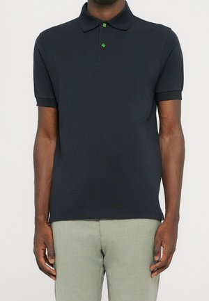 Uomo che indossa una polo nera a maniche corte con bottoni a forma di trifoglio verde e pantaloni grigio chiaro, in piedi contro uno sfondo semplice.