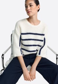 Fehér, bolyhos pulóver sötétkék csíkokkal, rövid ujjú és kerek nyakkivágással. A modell egy székben ül, sötét, testhez simuló nadrágot visel.