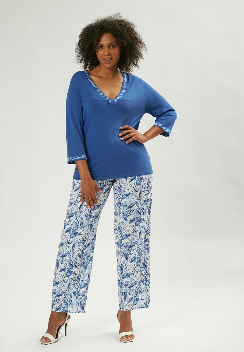 Top in maglia blu con scollo a V e bordatura decorata, abbinato a pantaloni larghi con stampa di foglie blu su sfondo bianco.