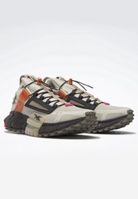 Zapatillas deportivas en beige y negro con suelas robustas, acentos en naranja y rosa, cordones negros con extremos en naranja y un diseño texturizado en la punta.