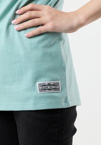 Mint green cotton t-shirt with a woven label displaying "DENIM FACTORY." Short sleeves, slightly loose fit, paired with black pants.