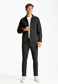 Sort skjorteglad jakke med brystlomme, kombineret med mørke, smalle bukser og hvide sneakers. Glat tekstur og skræddersyet design.