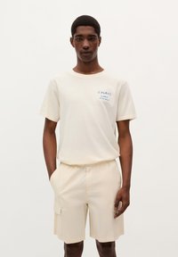 Tricou crem cu maneci scurte și text albastru, împreună cu pantaloni scurți bej tip cargo. Design simplu, textură moale și croială lejeră. Modelul este în poziție cu mâinile în buzunare.