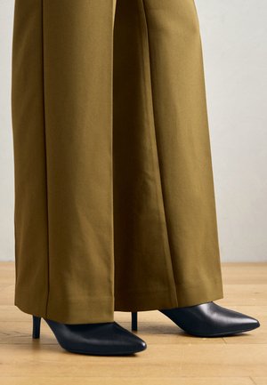 Schwarze spitze High Heels mit leichtem Glanz, getragen mit weit geschnittenen, senffarbenen Hosen. Glatte Textur und Stiletto-Absatzdesign.