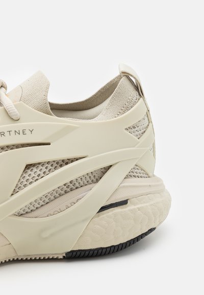 Beige athletic shoe with textured knit upper, curved overlay design, and a cushioned sole. Features a black rubber outsole and branding detail.