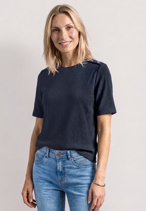 Femme blonde portant un haut texturé bleu marine à manches courtes et un jean bleu clair, souriante, avec les mains détendues sur les côtés, devant un fond uni.