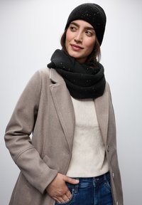 Gorro y bufanda de punto negros con detalle moteado, combinados con un abrigo beige claro y una parte superior crema, con un ajuste relajado y jeans azules.