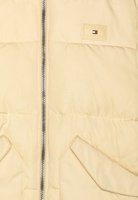 Beige dunjakke med lynlåslukning foran, to sidelommer og en logoemblem. Har glat tekstur og quiltet mønster.