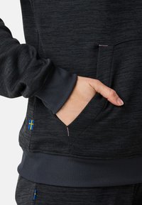 Felpa grigia scura con superficie texturizzata, dotata di tasca frontale e polsini a costine. Include un piccolo accento della bandiera svedese sul lato.