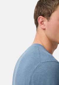 Nærbillede af en person iført en lyseblå striktrøje, der viser skulderen, halsen og øret mod en ensfarvet baggrund.