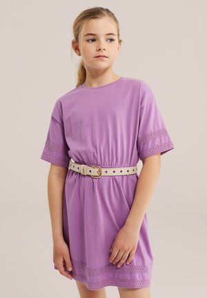 Junges Mädchen trägt ein fliederfarbenes Kleid mit Spitzenbesatz, tailliert mit einem beigen Gürtel, und steht vor einem schlichten hellen Hintergrund.