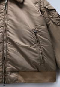 Brązowa ocieplana kurtka z kołnierzem z owczej skóry, zamkniętymi na zamek kieszeniami, prążkowanymi mankietami oraz gładką fakturą, z trwałym materiałem zewnętrznym.