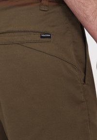 Braune Chino-Hosen mit glatter Textur, schräge Vordertaschen und einem schwarzen Volcom-Logoetikett, das auf der Gesäßtasche angenäht ist.
