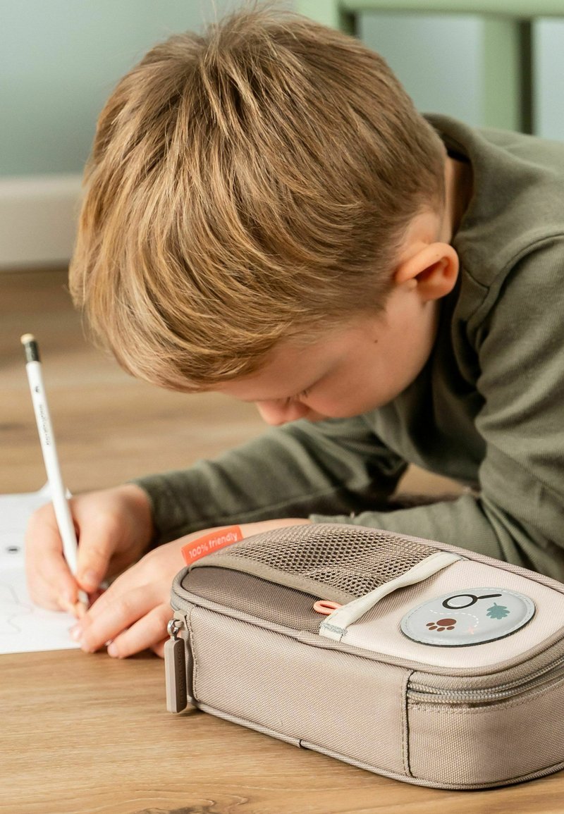 Enfant aux cheveux blonds allongé sur le sol écrivant au crayon à côté d'une trousse beige avec une poche en maille et un écusson circulaire.