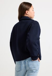 Mørkeblå ull-blandingsjakke med krage, senkede skuldre og en kort silhuett. Har minimalisk design og teksturert stoff.