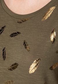 Camiseta de tela verde oliva con patrones de plumas metálicas doradas dispersas, con un cuello redondo y una superficie suave y ligeramente texturizada.