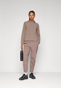 Bruine sportjack en broek met elastische manchetten, gecombineerd met donkere sneakers en een zwarte waterfles. Glad, lichtgewicht materiaal.