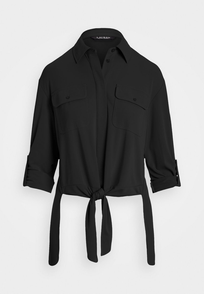 Chemise noire à manches longues avec poignets retournés, deux poches poitrine boutonnées, fermeture boutonnée devant et un lien à nouer à l’ourlet.