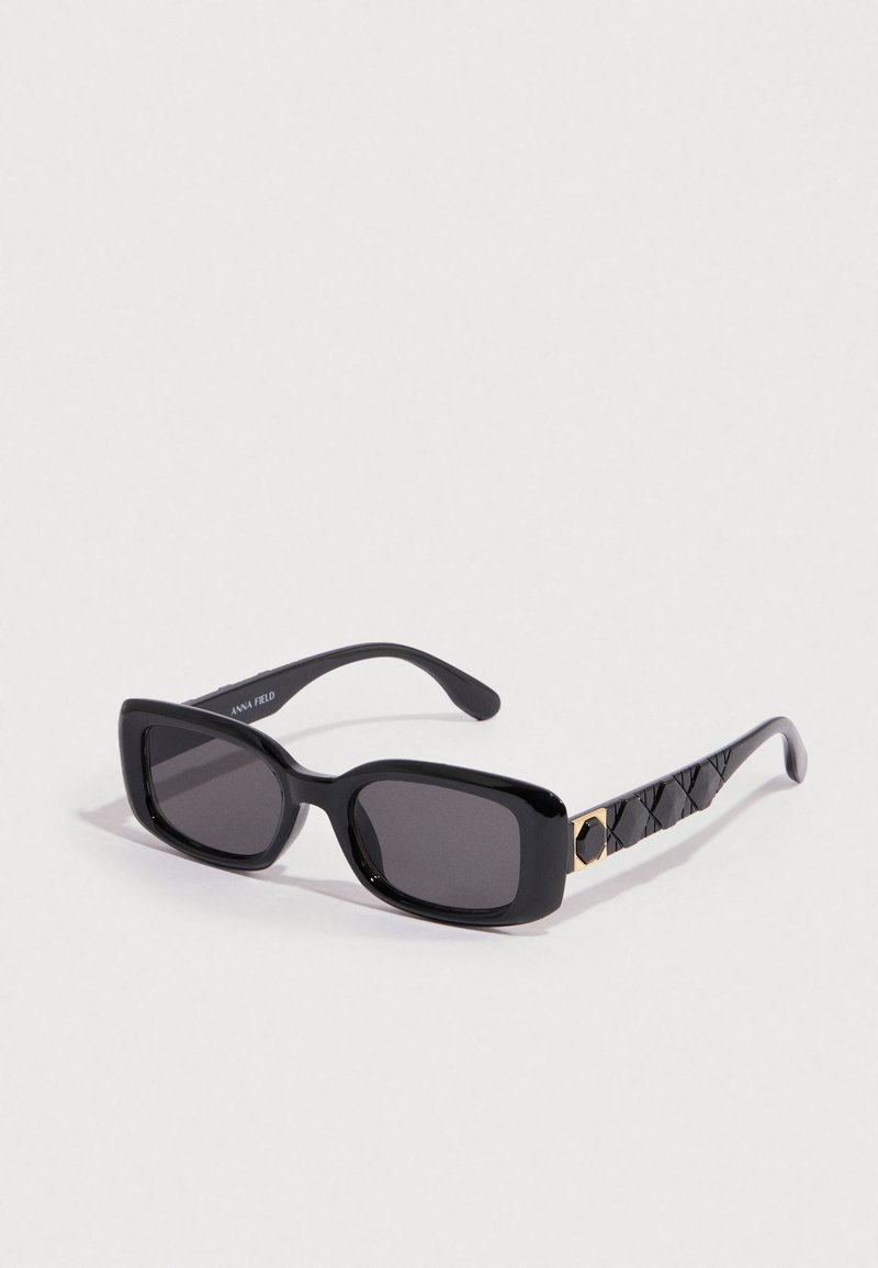 Gafas de sol rectangulares negras con lentes oscuras. La montura tiene un acabado brillante y un acento decorativo de patrón negro y dorado en la patilla.