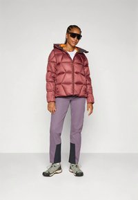 Casaco puffer marrom com capuz e forro laranja, combinado com calças lilás claras com detalhes pretos e botas de caminhada cinzas.