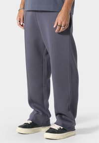 Pantalons de survêtement gris surdimensionnés avec une texture douce, présentant un design à jambe droite et des coutures contrastantes. Portés avec des baskets noires.