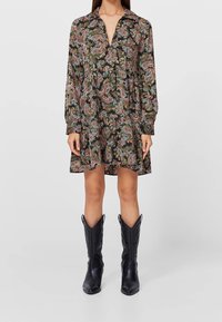 Mujer vestida con un vestido de manga larga con un patrón de paisley y botas vaqueras negras hasta medio pantorrilla, de pie frente a un fondo gris liso.