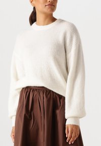 Frau trägt einen weißen, gerippten Strickpullover mit langen Ärmeln und einen braunen, hoch taillierten, gerafften Rock, steht vor einem schlichten Hintergrund.