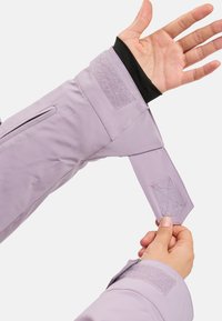 Light lavender jacket sleeve with black inner lining, featuring a Velcro strap and a side zipper pocket. Smooth, textured fabric.