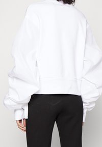 Vit oversized sweatshirt med voluminösa ärmar, ribbad nederkant och slät textur. Kombinerad med svarta tajta byxor. Bakifrån visad.