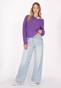 Vijolična oversized pletena pulover s rebrastimi manšetami, v kombinaciji s svetlo modrimi širokimi hlačami. Model nosi bele in metine superge.