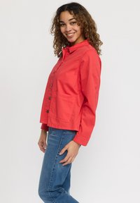 Röd knäppt jacka med krage, lång ärm och en framficka. Har en lätt och slät textur. Stylad med blå jeans.