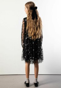 Czarna sukienka z tiulu z wzorem srebrnych gwiazdek, długimi rękawami, warstwowym krojem i czarną aksamitną kokardą z tyłu. Czarne buty i podkolanówki.