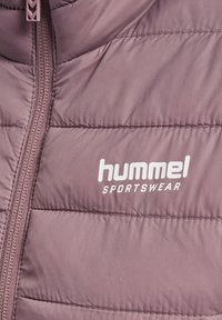 Gros plan d'une veste matelassée mauve avec une fermeture éclair verticale et le logo blanc "hummel SPORTSWEAR" sur la partie droite de la poitrine.