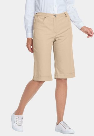 Shorts en coton beige arrivant au genou avec des revers et des poches avant, associés à des baskets blanches. Texture de tissu lisse, coupe décontractée.