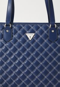 Granatowa torba z pikowanego materiału z białym haftem w diamenty, z okrągłą srebrną blaszką z logo w kształcie trójkąta oraz dopasowanymi uchwytami. Gładki materiał syntetyczny.