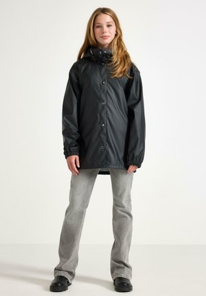 Giovane donna che indossa un impermeabile nero con cappuccio, jeans grigi chiari e stivali neri, in piedi davanti a uno sfondo bianco uniforme.
