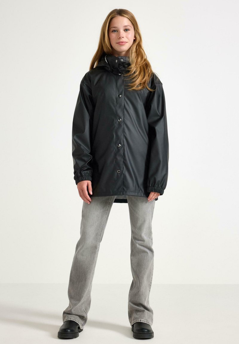 Giovane donna che indossa un impermeabile nero con cappuccio, jeans grigi chiari e stivali neri, in piedi davanti a uno sfondo bianco uniforme.