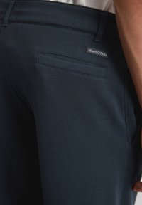Pantalons bleu marine foncé en tissu texturé. Présentent une poche arrière avec une petite étiquette de marque Marc O'Polo. La ceinture est lisse et ajustée.