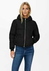 Veste matelassée noire avec capuche, fermeture éclair à l'avant et poignets élastiques. Dotée d'une texture matelassée verticale et de poches latérales. Portée avec un jean bleu.