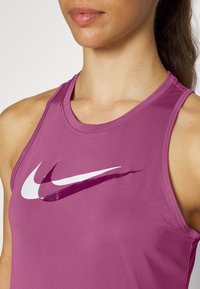 Fioletowy sportowy tank top z okrągłym dekoltem i szerokimi wycięciami na ramionach. Posiada duże białe logo Nike na klatce piersiowej, gładka tekstura materiału.
