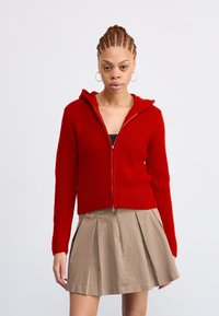 Rode gebreide hoodie met rits en capuchon, met ribbelstructuur, gecombineerd met een beige geplooide rok met een klein logo.