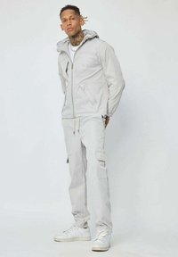 Veste à zip gris clair avec capuche, dotée de deux poches avant, associée à un pantalon cargo assorti et des baskets blanches. Texture lisse.
